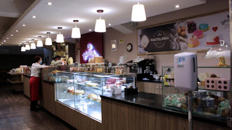 Interior de una pastelería moderna y bien iluminada, con vitrinas de vidrio que exhiben una variedad de postres y productos de repostería. El espacio cuenta con un largo mostrador de madera, lámparas colgantes de luz blanca y paredes decoradas con imágenes de dulces.