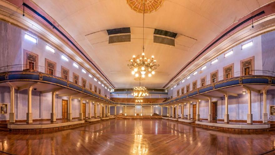 La imagen muestra el interior de un teatro o salón amplio y elegante, con un piso de madera brillante y balcones laterales de estilo clásico. Grandes lámparas colgantes iluminan el espacio, resaltando los detalles arquitectónicos y creando una atmósfera cálida y majestuosa.