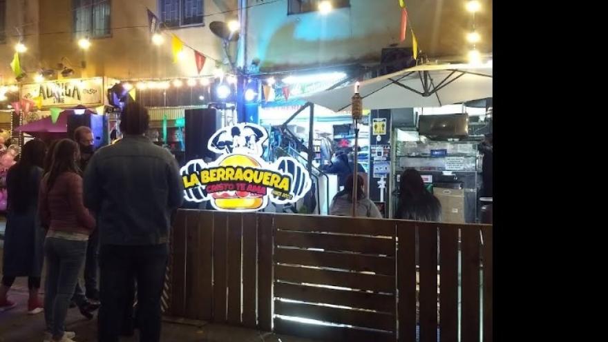 Escena nocturna en una zona gastronómica al aire libre, con varias personas reunidas frente a un puesto de comida llamado “La Berraquera”. El lugar está rodeado por una cerca de madera y decorado con luces colgantes y banderines de colores.