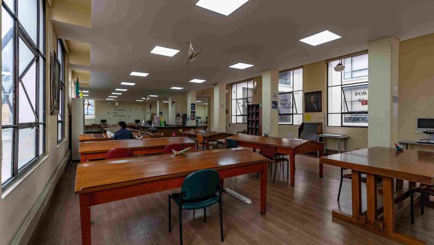 La imagen muestra una sala de lectura amplia y ordenada, con mesas de madera y sillas distribuidas en filas. La luz natural entra por grandes ventanas, complementada por iluminación en el techo.