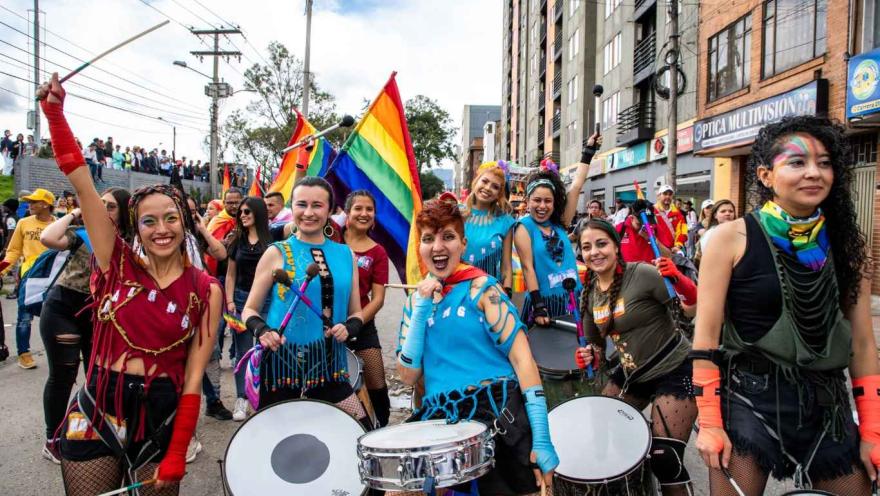  Grupo de personas marchando en un desfile del orgullo LGBTI+ por una calle urbana, tocando tambores y sonriendo; llevan vestuarios coloridos, maquillaje llamativo y banderas arcoíris, mientras una multitud los acompaña a ambos lados de la vía.