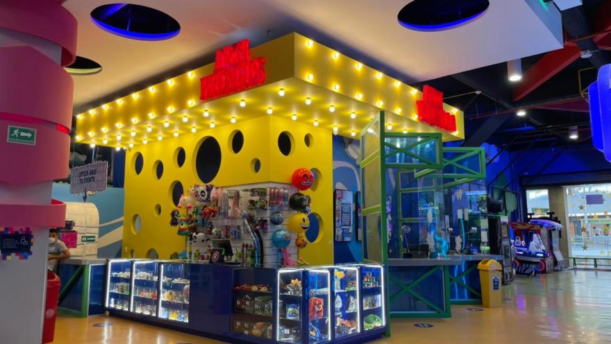 Mostrador de premios con vitrinas iluminadas en un parque de entretenimiento interior, rodeado de juegos y decoración colorida.