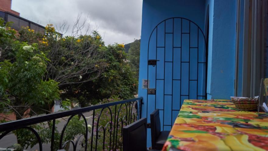 La imagen muestra un balcón acogedor con paredes azules y una mesa cubierta con un mantel colorido. Una baranda de hierro enmarca la vista hacia árboles y una calle tranquila, bajo un cielo nublado. El espacio transmite calma y un ambiente íntimo al aire libre.