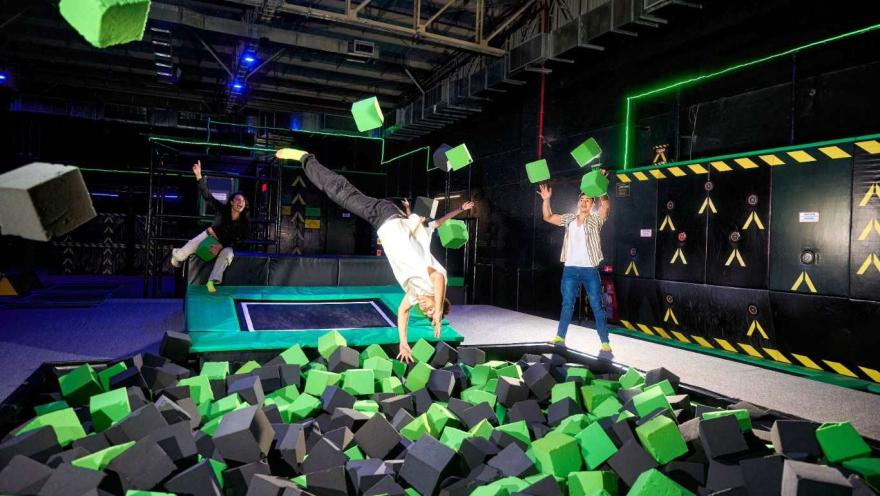 Personas saltan y juegan en una piscina de cubos de espuma, mientras una de ellas realiza una voltereta en un parque de trampolines interior.