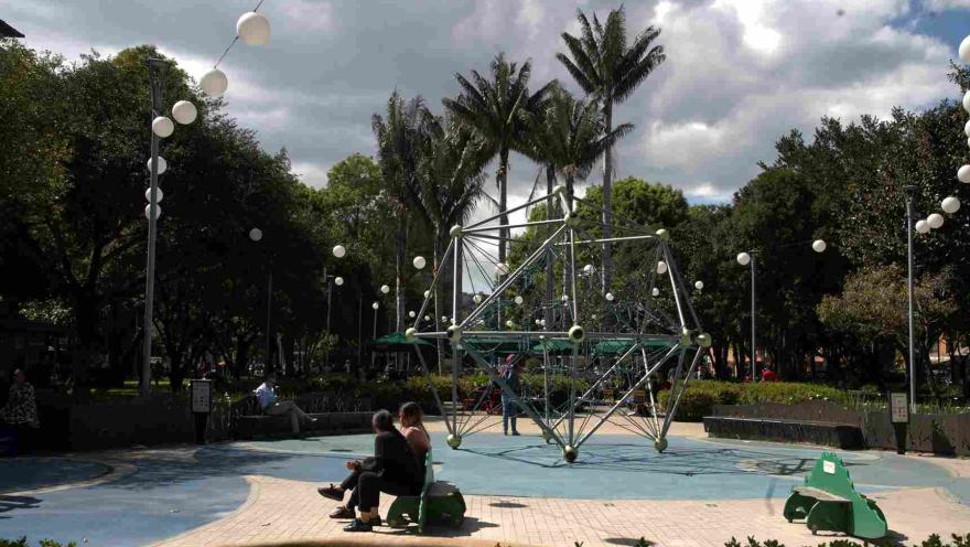 Área de juegos con una estructura metálica para escalar, rodeada de árboles y personas sentadas en bancas. 
