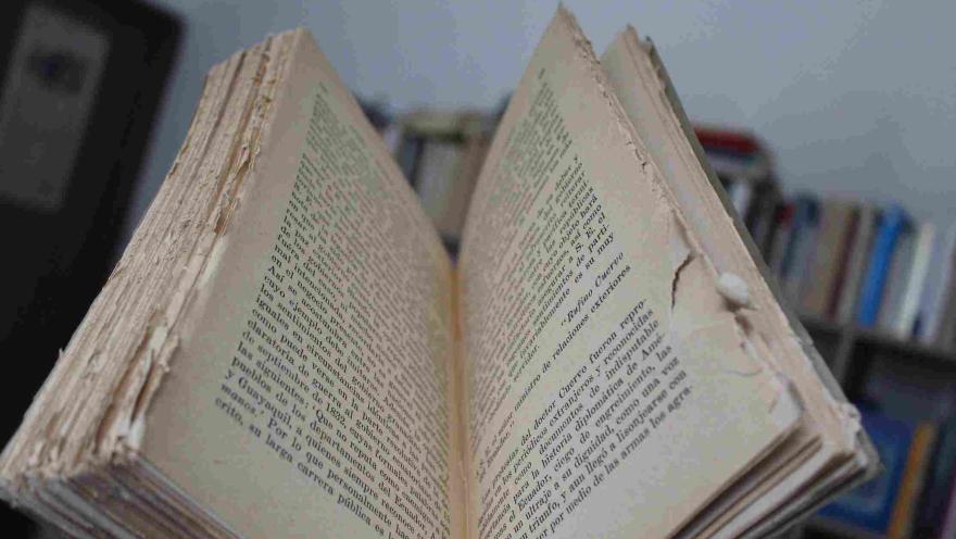  Libro antiguo abierto con páginas desgastadas y texto visible, sobre fondo desenfadado de estantería.