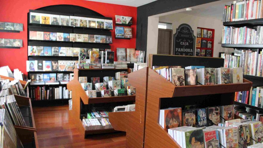  Interior de librería con estanterías de madera llenas de libros y cómics, con pared roja y exhibidores organizados.