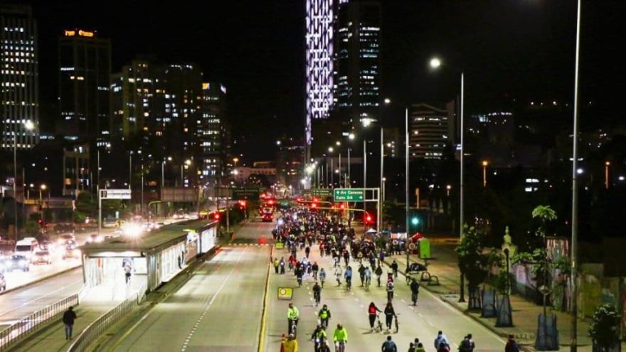 Ciclistas recorren una vía principal de Bogotá durante la noche, con edificios iluminados y tráfico alrededor.