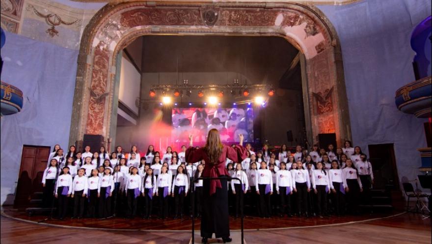 La imagen muestra un coro numeroso de niños y jóvenes sobre un escenario, dirigidos por una mujer ubicada al frente. La escena está iluminada con luces cálidas y de colores, mientras una pantalla al fondo complementa la presentación.