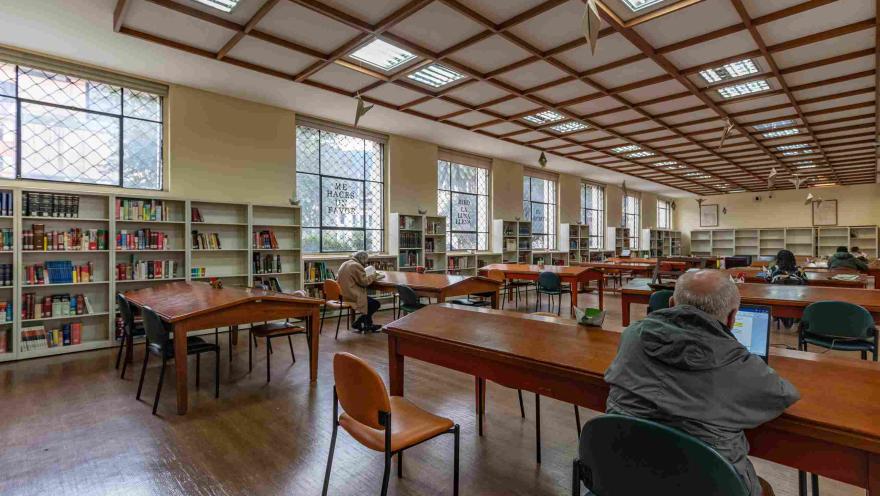 La imagen muestra una sala de lectura amplia y bien iluminada, con mesas de madera y estanterías llenas de libros a lo largo de las paredes. Varias personas leen o trabajan en silencio, creando un ambiente tranquilo y concentrado. La luz natural que entra por las ventanas y el techo geométrico aportan calidez y orden al espacio.