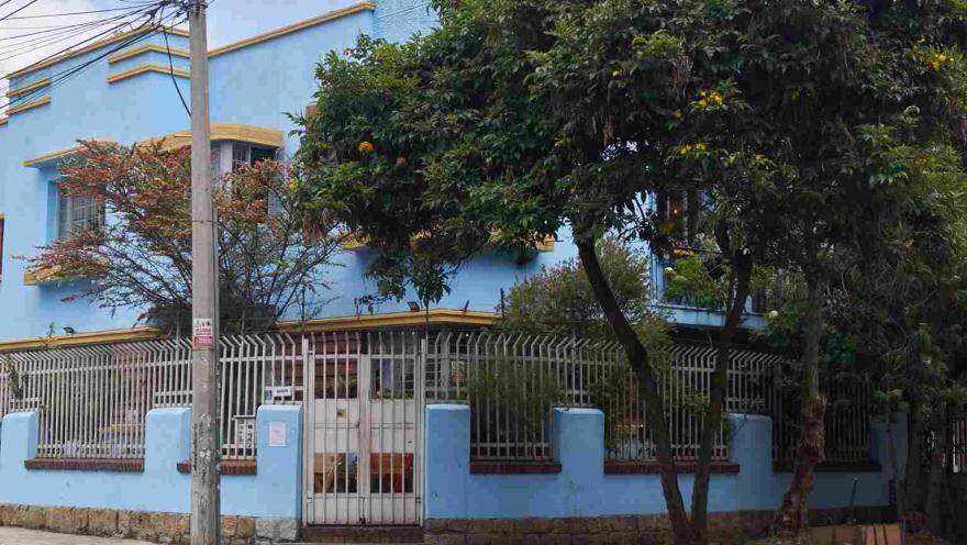 La imagen muestra una casa de dos pisos pintada en tonos azul claro con detalles amarillos, rodeada por una reja blanca. Al frente hay árboles que aportan sombra y vegetación, mientras un poste y cables eléctricos evidencian el entorno urbano. La escena transmite tranquilidad en un barrio residencial.