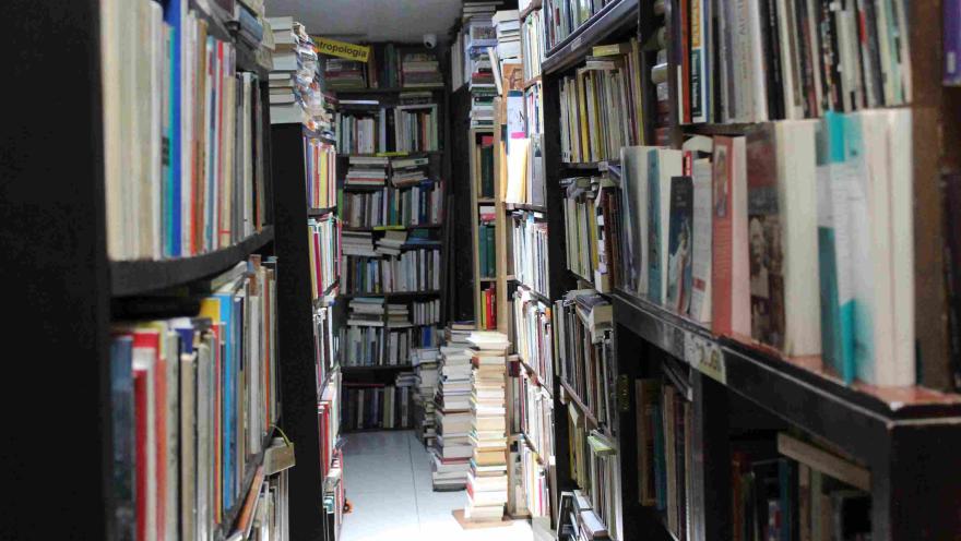Pasillo estrecho de librería con estanterías llenas de libros y pilas en el suelo.