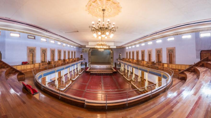 La imagen muestra el interior de un teatro de estilo clásico visto desde un nivel superior, con balcones curvos y un escenario al fondo. El piso de madera y las lámparas ornamentales aportan calidez, mientras la disposición circular del espacio resalta su elegancia y amplitud.