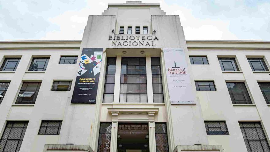 La imagen muestra la fachada de un edificio institucional de estilo sobrio y simétrico, identificado como la Biblioteca Nacional. Su diseño en tonos claros y líneas verticales resalta la entrada principal, flanqueada por grandes ventanales y banners informativos.