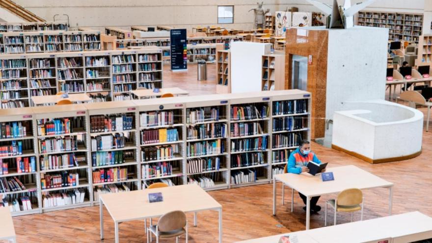  Interior de biblioteca amplia con estanterías llenas de libros y una persona leyendo en una mesa.