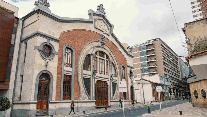 La imagen muestra la fachada de un edificio histórico de estilo ornamental, con detalles en ladrillo y piedra que destacan en el entorno urbano. Su diseño elegante contrasta con construcciones modernas cercanas, mientras algunas personas caminan por la calle, aportando vida a la escena bajo un cielo nublado.