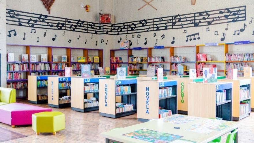 Interior de una biblioteca pública infantil, con estanterías bajas llenas de libros organizados por categorías como “novela” y “cuento”. El espacio es colorido, con muebles en tonos vivos, mesas de lectura y pufs, y una pared decorada con un gran pentagrama y notas musicales que recorre todo el salón, creando un ambiente lúdico y acogedor para niños.