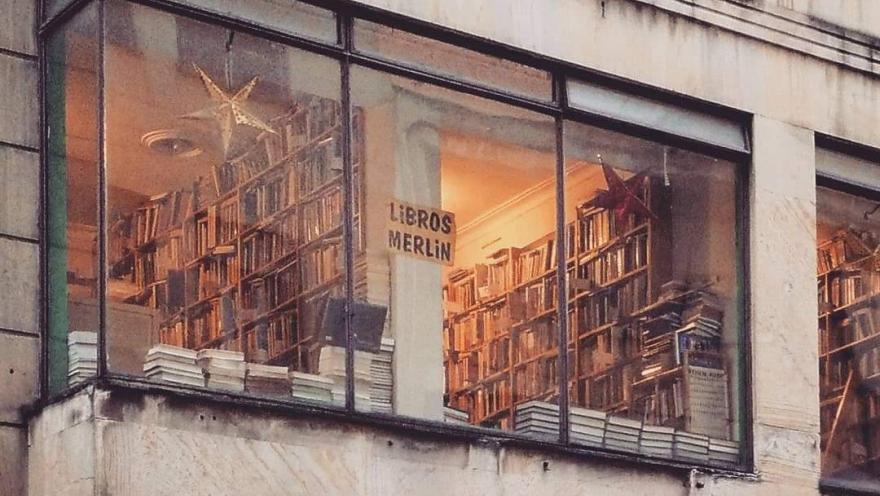 Ventana exterior desde la que se observa la Librería Merlín con estanterías llenas de libros, iluminadas cálidamente desde el interior.