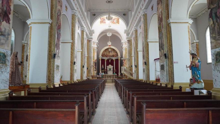 Interior de una iglesia con pasillo central y bancas de madera, columnas decoradas y un altar al fondo enmarcado por arcos y pinturas religiosas.