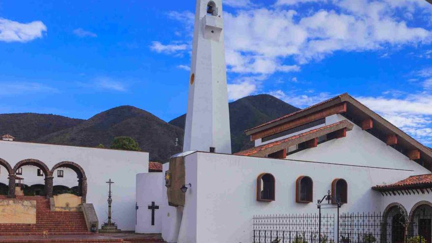 Iglesia de arquitectura blanca con campanario alto, rodeada de montañas y cielo azul, con una plaza de piedra en primer plano.