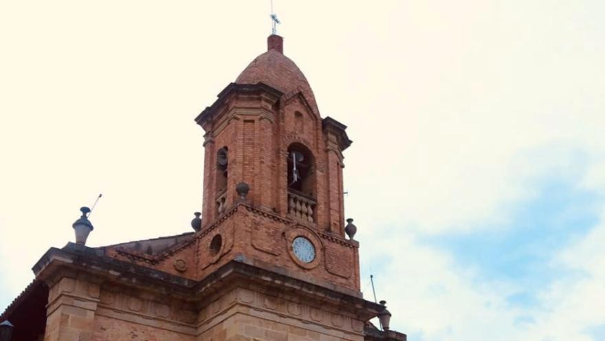 Torre de una iglesia de ladrillo con campanario y reloj, coronada por una cruz, contra un cielo claro con nubes.