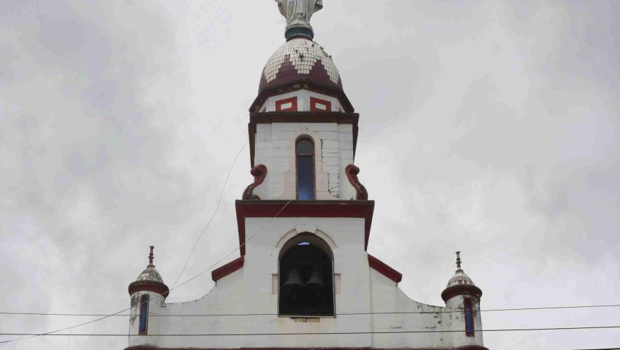  Torre de iglesia blanca con detalles rojos, coronada por una estatua de Cristo con los brazos abiertos, contra un cielo nublado.