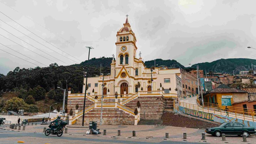  Plano general de la fachada de una iglesia color marrón claro, tras la cual hay montañas. Al frente hay una calle con un automovil estacionado y un motociclista.