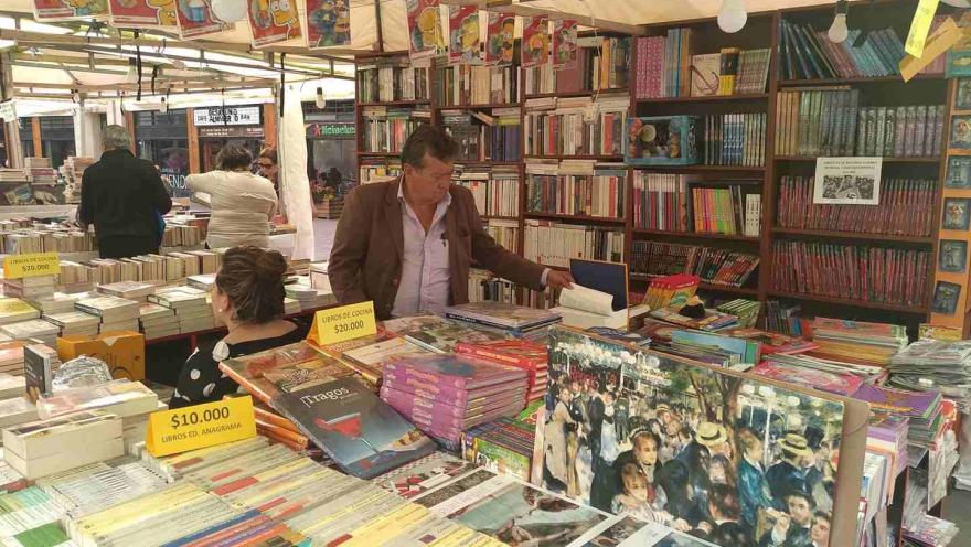 Puesto de venta de libros en un mercado, con estanterías llenas y personas explorando y comprando entre títulos variados.