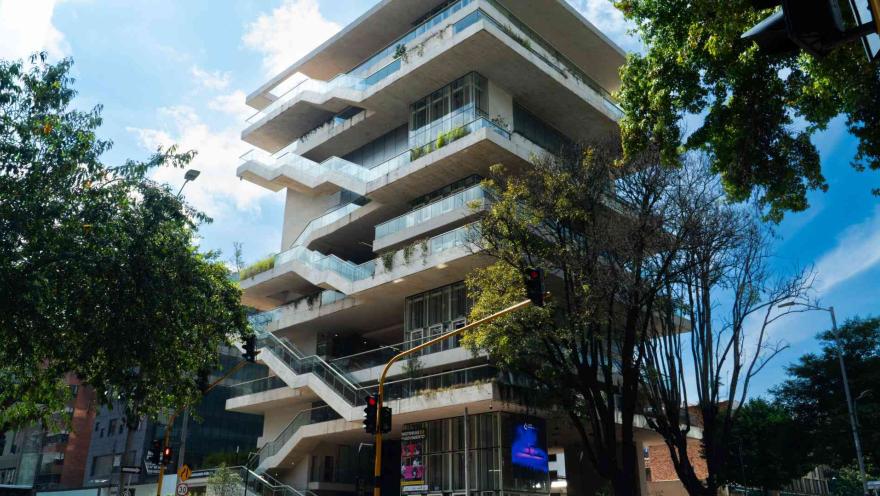  Edificio moderno de varios niveles con terrazas escalonadas y amplios ventanales, rodeado de árboles en una esquina urbana bajo cielo despejado.