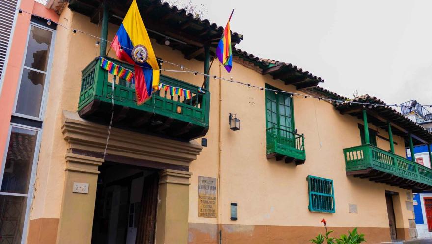 Fachada de casa colonial con balcones de madera pintados de verde, decorada con banderas de Colombia y colores del orgullo, en una calle de estilo tradicional.