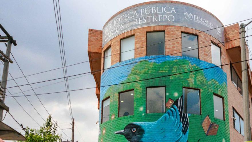Fachada de la Biblioteca Pública Carlos E. Restrepo con un mural colorido de un ave y libros, que resalta en el entorno urbano bajo un cielo nublado.