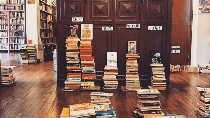  Interior de librería con pilas de libros organizadas por temas sobre piso de madera y estanterías al fondo.
