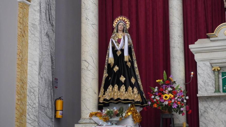 Escultura de la Virgen vestida de negro con detalles dorados, ubicada entre columnas y un fondo de cortinas rojas, acompañada de flores y velas.
