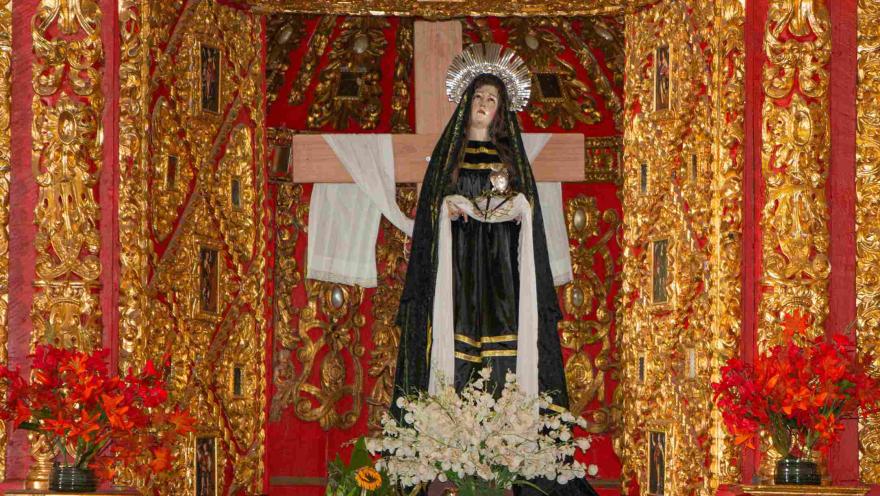 Altar religioso decorado en dorado y rojo, con una escultura de la Virgen de los Dolores frente a una cruz, rodeada de flores y ornamentos barrocos.