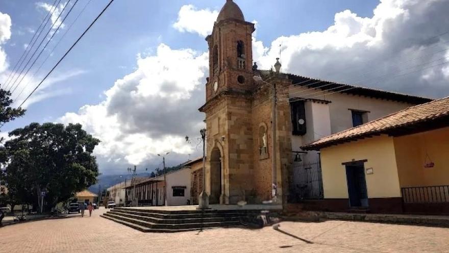 Iglesia de pueblo con torre de piedra y campanario, ubicada junto a una plaza amplia, con casas coloniales y cielo parcialmente nublado.
