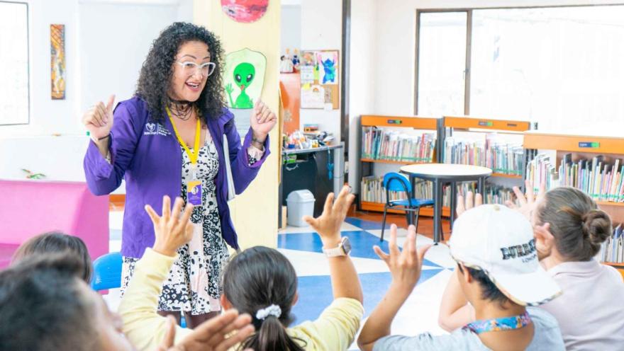 Mediadora en una biblioteca interactúa con un grupo de niños que levantan la mano, en un espacio colorido que fomenta la lectura y el aprendizaje.