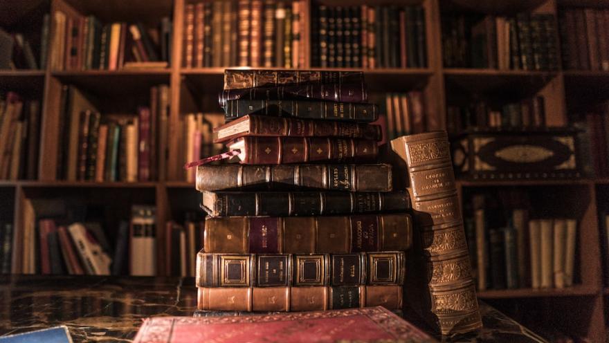 Pila de libros antiguos sobre una mesa, con estanterías llenas de volúmenes al fondo en una biblioteca cálida.