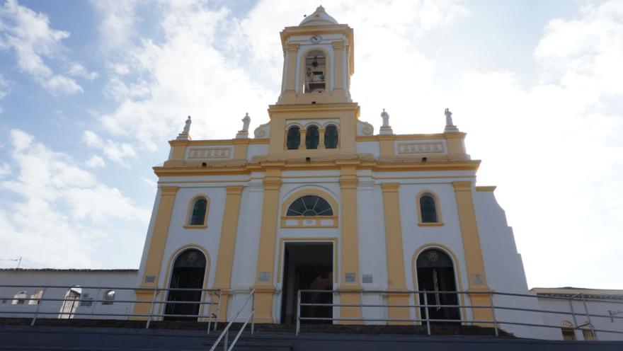 Fachada de una iglesia de estilo colonial en tonos blanco y amarillo, con torre central y escalinatas, vista desde abajo contra un cielo parcialmente nublado.
