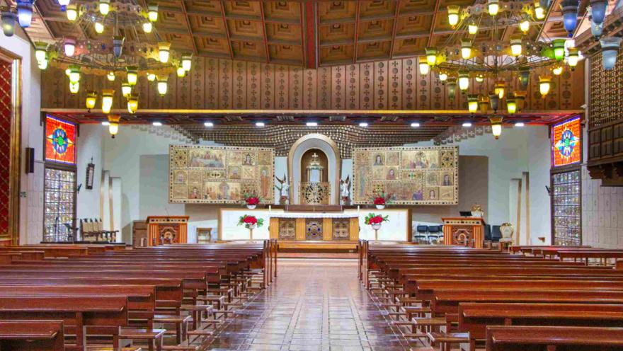  Interior de una iglesia con bancas de madera alineadas, lámparas colgantes y un altar decorado con imágenes religiosas y flores al fondo.