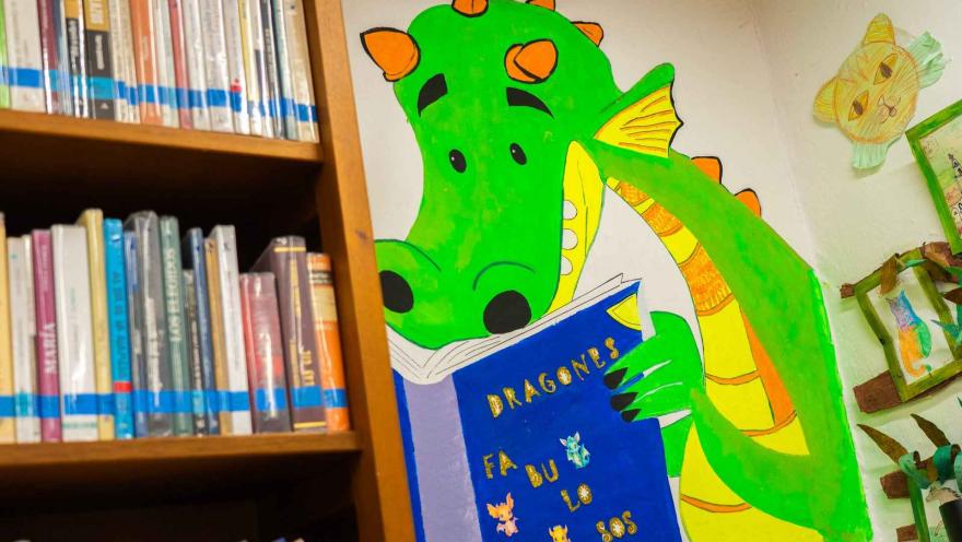 Mural colorido de un dragón verde leyendo un libro en una biblioteca, junto a estanterías llenas de libros que aportan un ambiente creativo y educativo.