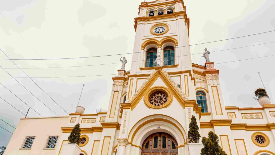 Primer plano de la fachada de una iglesia color marrón claro con toques de color amarillo. Hay algunos pinos pequeños a los lados.