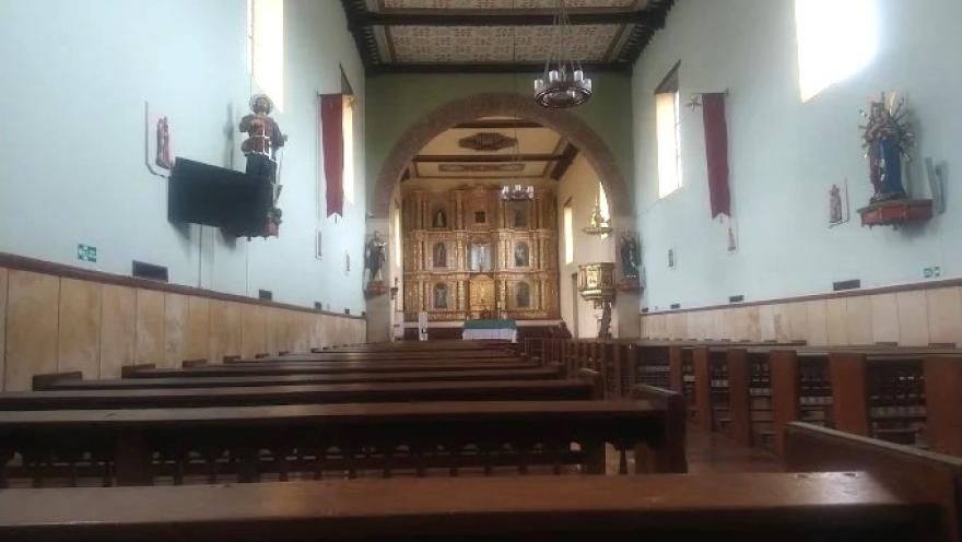 Interior de una iglesia con bancas de madera, paredes blancas y un altar dorado al fondo, iluminado por luz natural que entra por ventanas laterales.