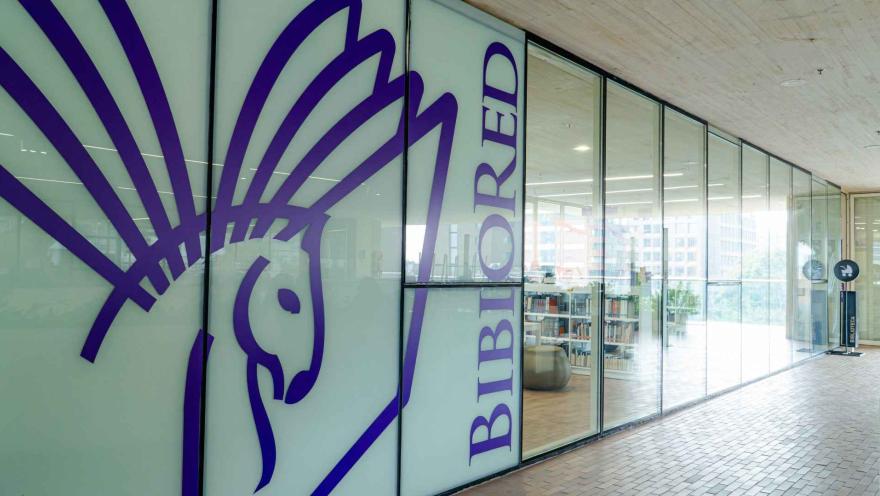 Fachada de biblioteca con paredes de vidrio y un diseño gráfico de colibrí en color púrpura, que identifica el espacio como un lugar moderno y cultural.