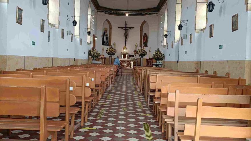 Interior de una iglesia con bancas de madera alineadas, piso geométrico y un altar al fondo con una cruz y arreglos florales.