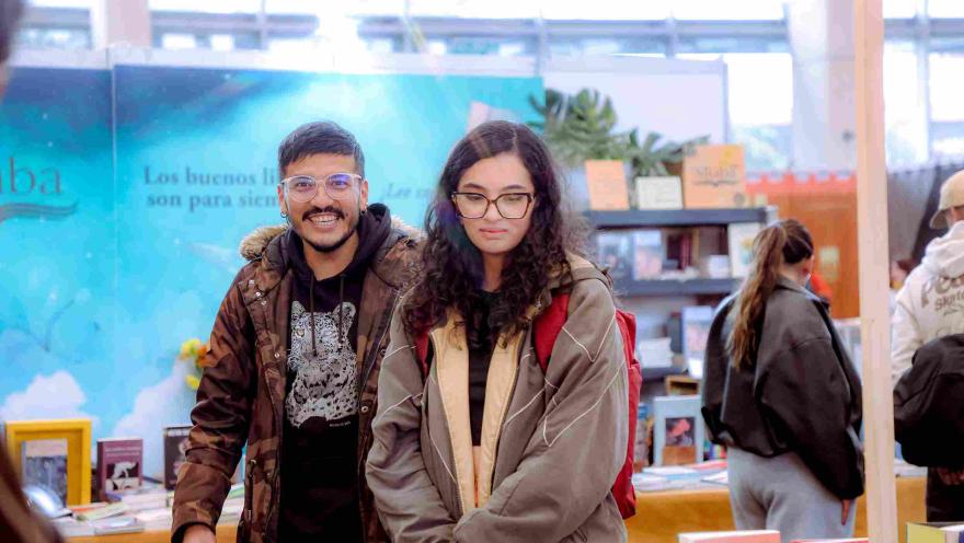 Dos visitantes posan y recorren un stand de libros en una feria literaria, en medio de un ambiente cultural lleno de lectores, editoriales y actividades alrededor de la lectura.