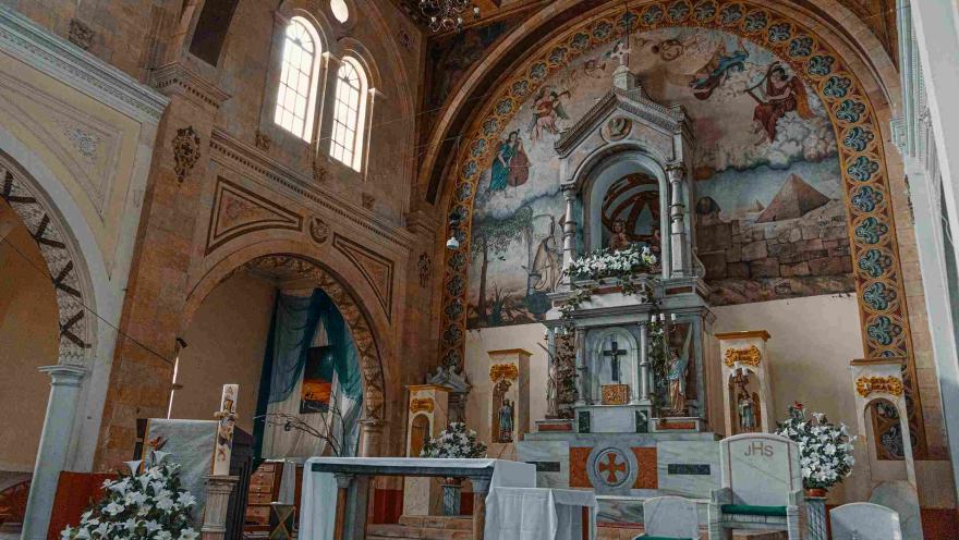  Interior de iglesia en la que sobresale un atrio con mantel blanco y arreglos florales con flores blancas.