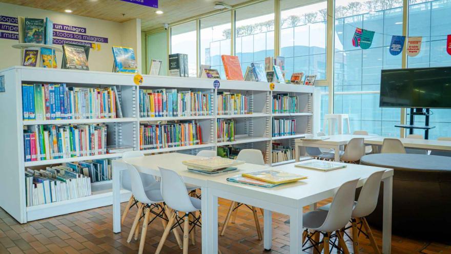  Sala de biblioteca con estanterías llenas de libros y mesas de estudio, iluminada por grandes ventanales que crean un ambiente cómodo para la lectura.