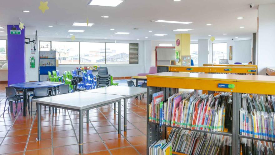 Sala infantil de biblioteca con estanterías de libros, mesas de trabajo y sillas de colores, en un espacio amplio y luminoso para la lectura y el aprendizaje.