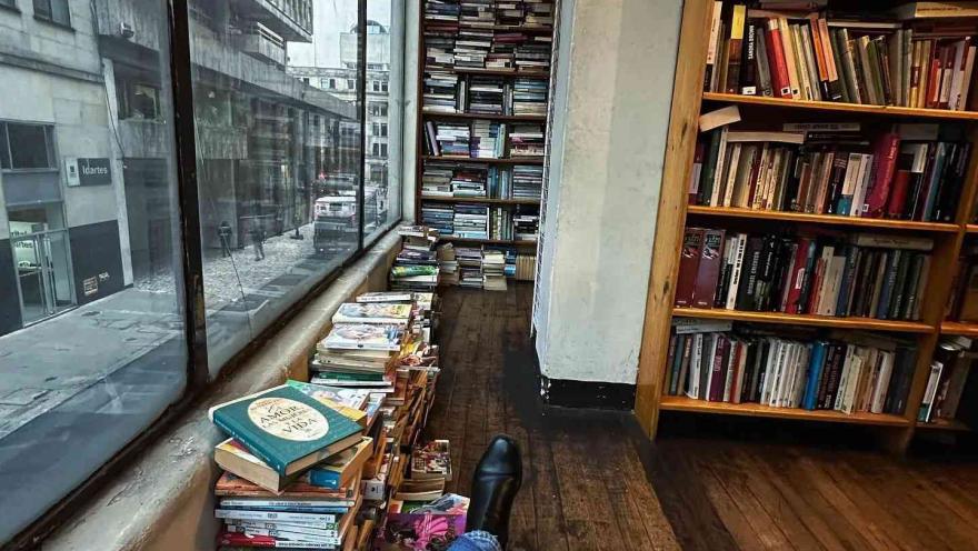 Vista desde una librería con estanterías y pilas de libros junto a una ventana, mientras una persona descansa mirando hacia la calle.