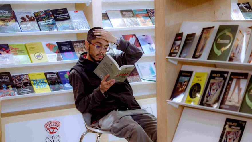 Un visitante lee concentrado un libro mientras descansa frente a estanterías llenas de publicaciones y novedades editoriales en una feria literaria.